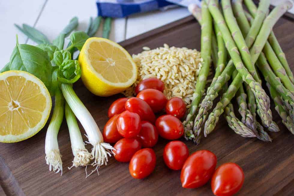 A board topped with asparagus, tomatoes, orzo, green onions and a half of a lemon.