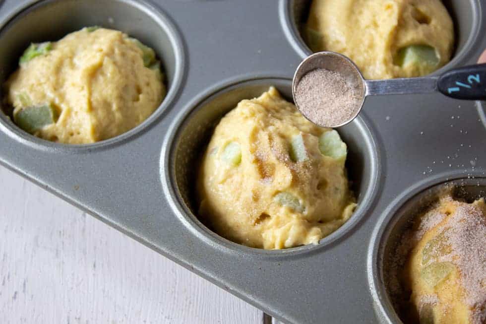 A muffin tin filled with rhubarb muffin batter being topped with cinnamon and sugar.