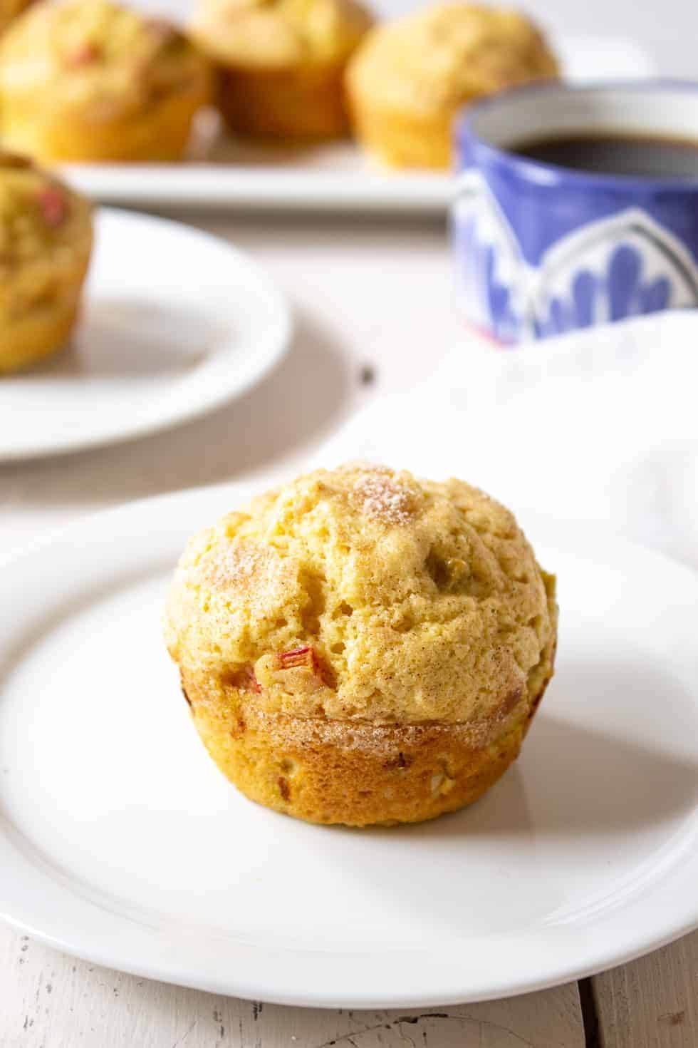 A rhubarb muffin on a small white plate with more muffins in the background. 
