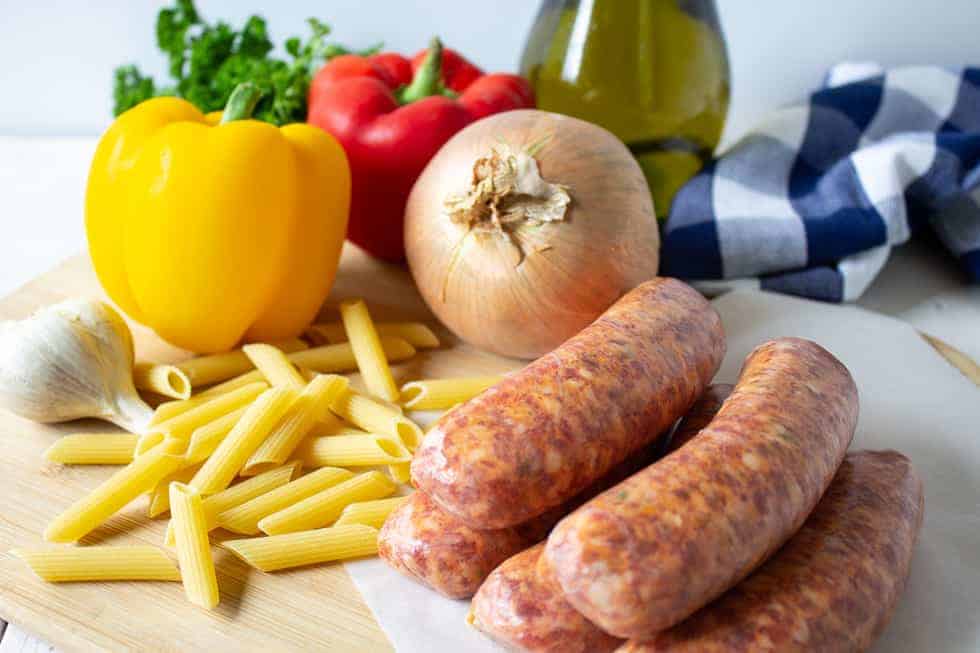 An arrangement of uncooked sausage, pasta and fresh onions and bell peppers on a wooden board.