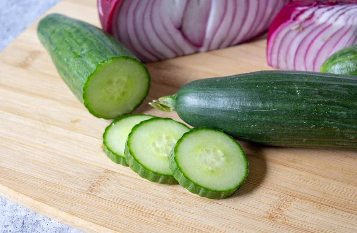 An English cucumber sliced on a cutting board with a red onion on the board too. 