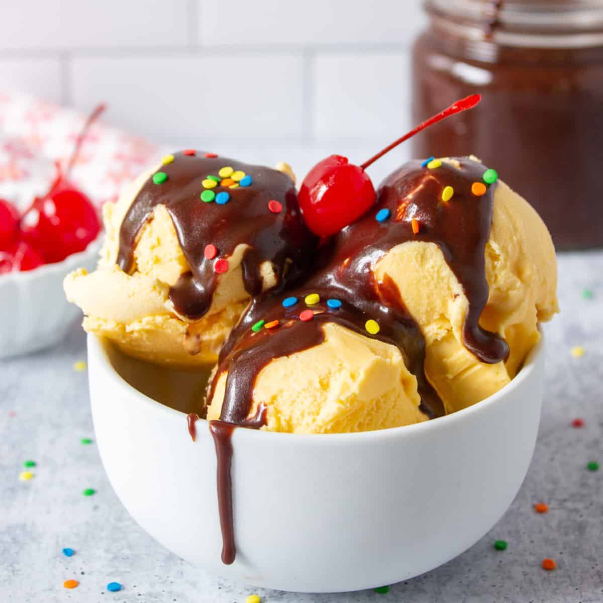 Hot fudge sauce dripping on top of vanilla ice cream topped with colored sprinkles and a cherry.