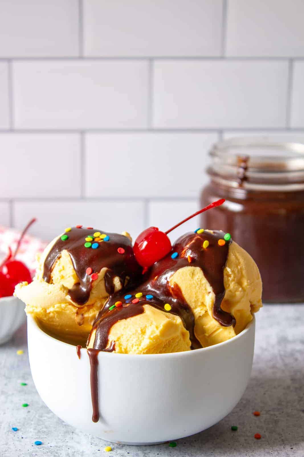 A white bowl with scoops of ice cream topped with hot fudge, sprinkles and a cherry. 