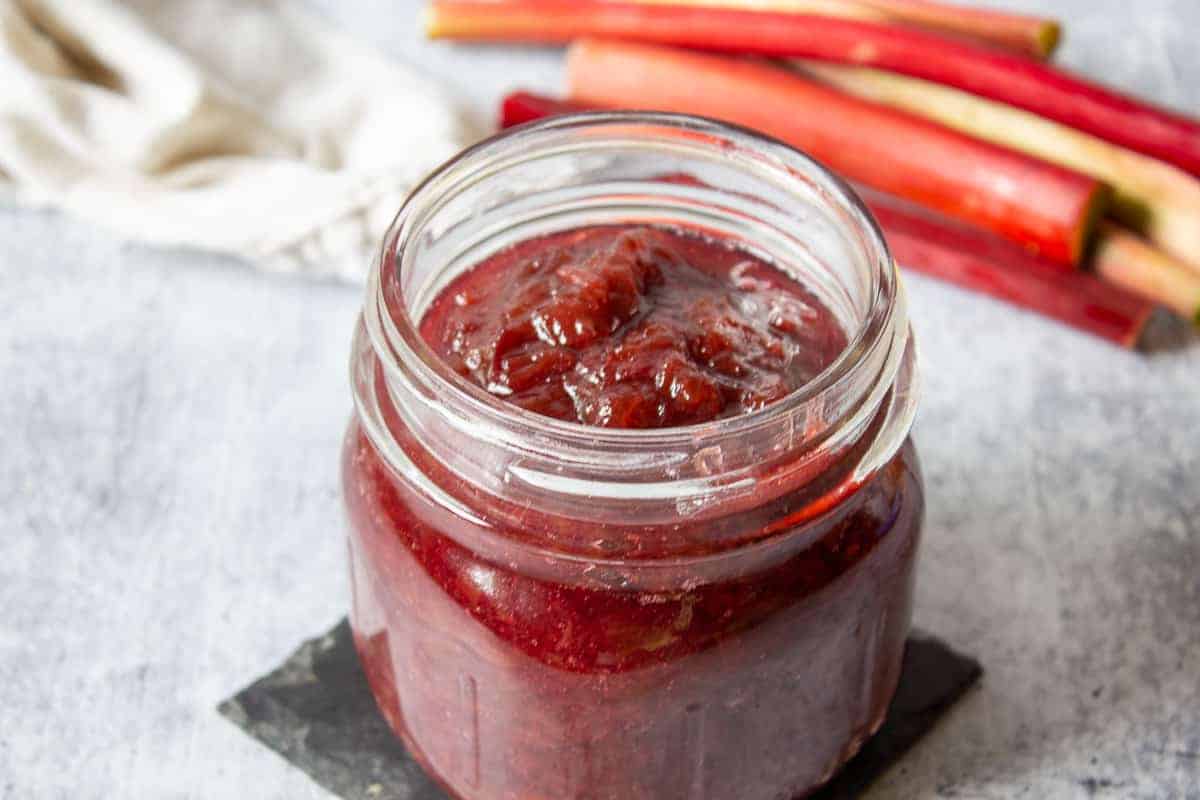 Homemade rhubarb jam in a glass canning jar with stalks of rhubarb in the background. 