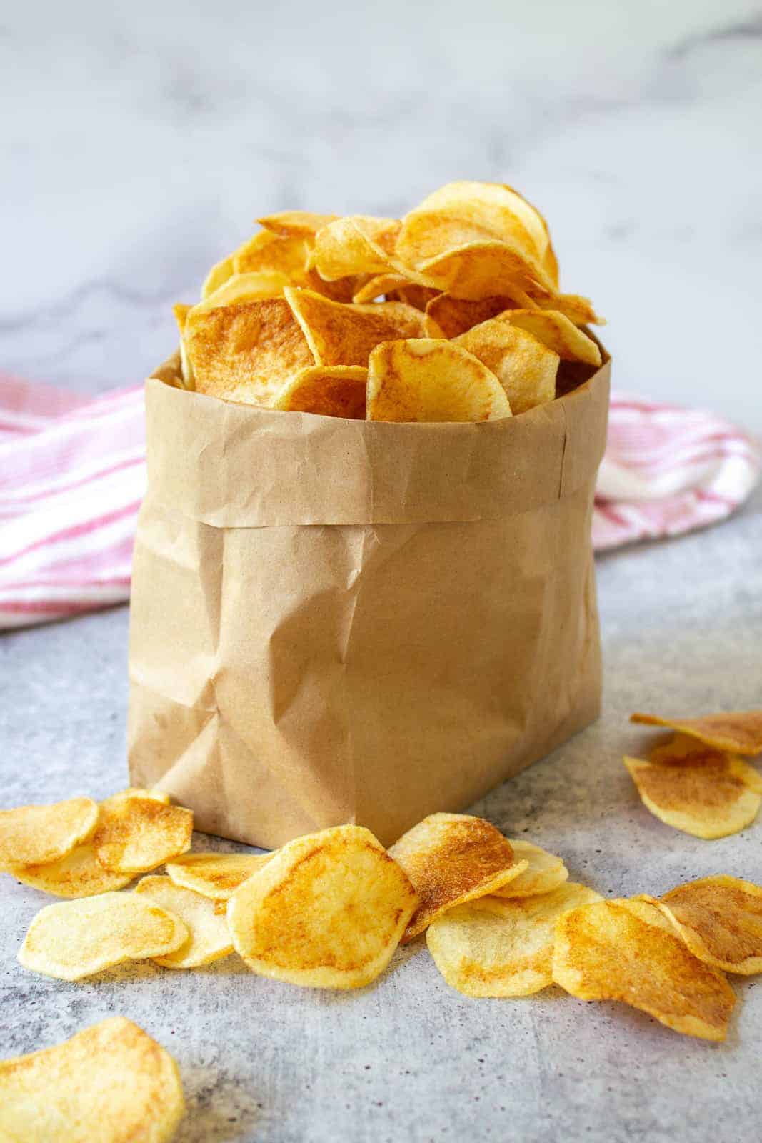 Potato chips stacked in a brown paper bag with a few chips sprinkled outside the bag.