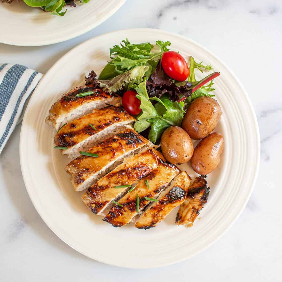 Sliced chicken breast on a white plate along with small red potatoes and a green salad.