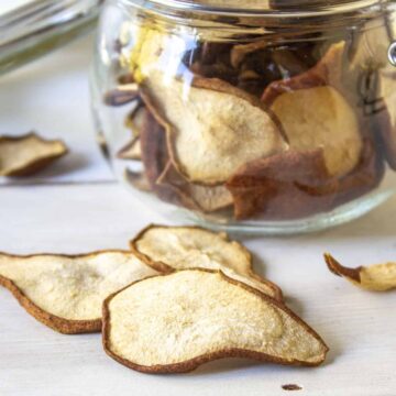 Dried pears on a board with a glass jar filled with pear chips behind them.