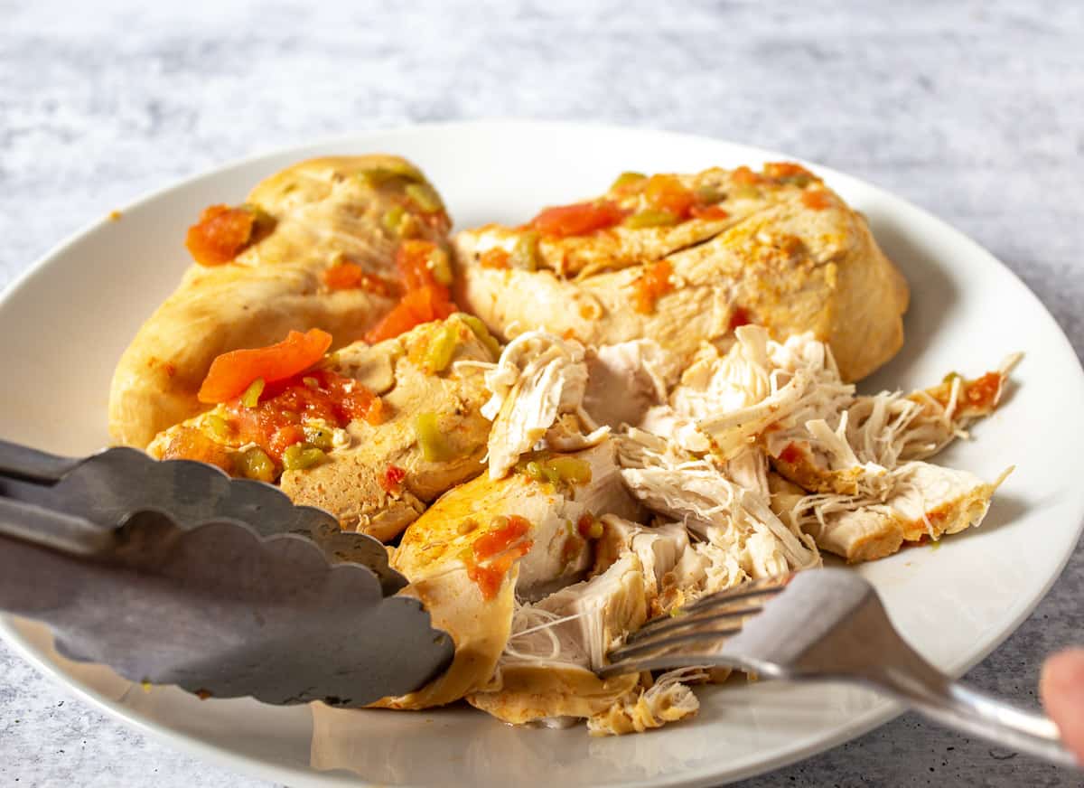 Chicken breasts on a plate being shredded with a fork and a pair of tongs.