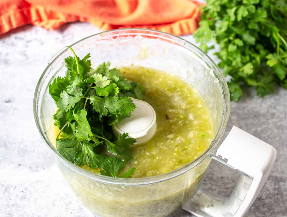 Fresh cilantro added to a food processor bowl filled with salsa verde.