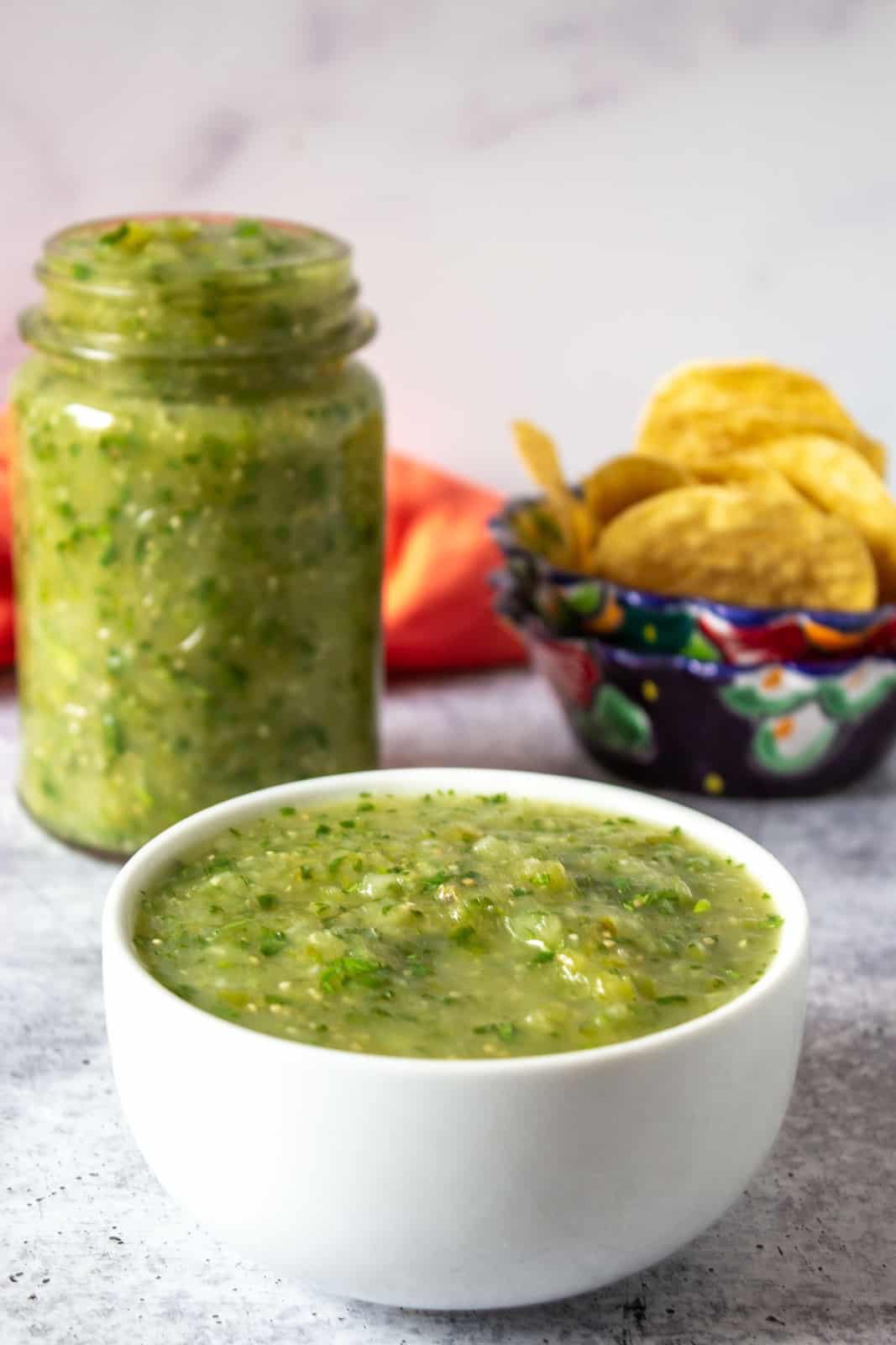 A bowlful of green salsa with a jar filled with salsa in the background.