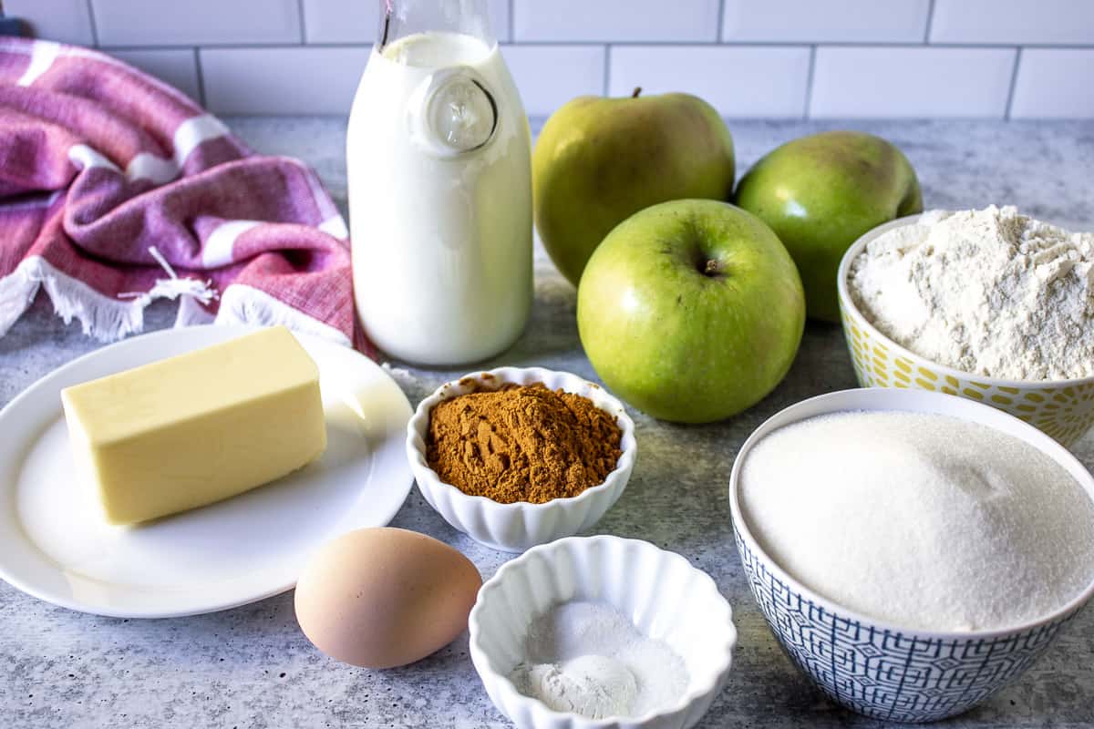 A variety of ingredients including apples, milk, butter, sugar and flour on a board.