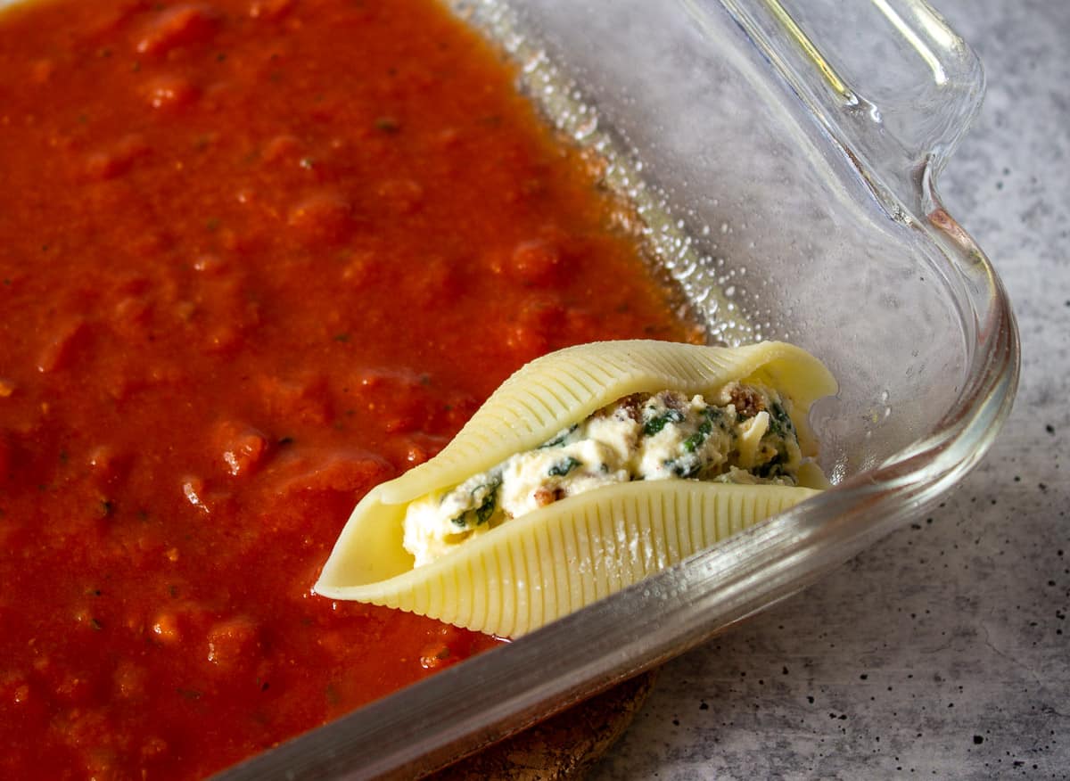 A glass dish with a layer of marinara sauce and one filled pasta shell in the dish.