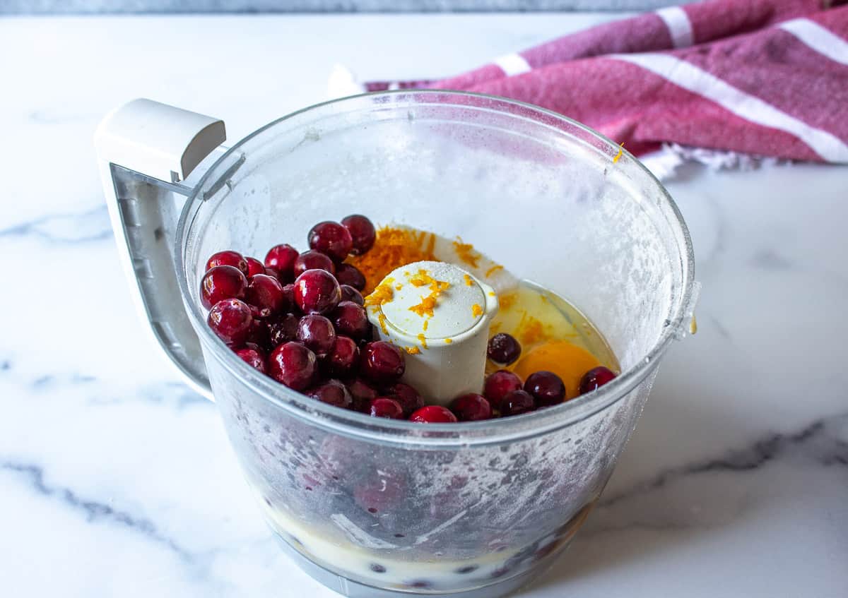 A food processor bowl with fresh cranberries, orange peel and an egg.