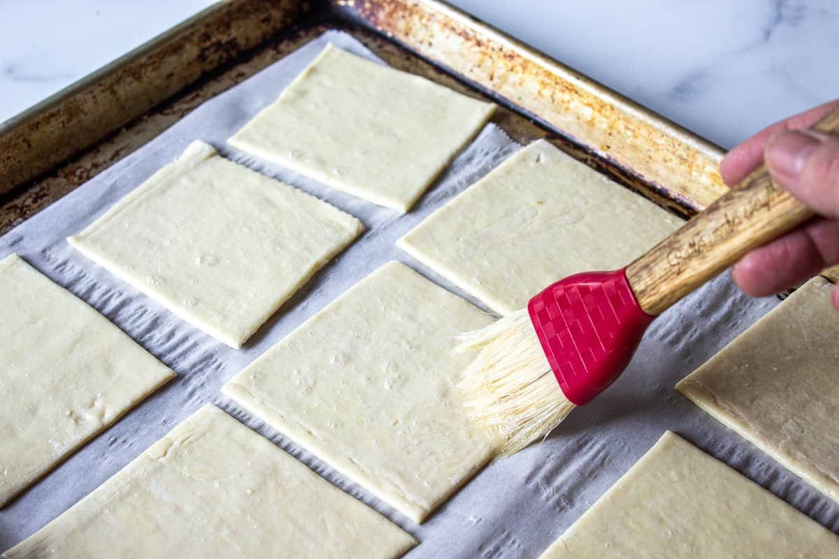 Puff pastry squares on a baking sheet being brushed with a small pastry brush.