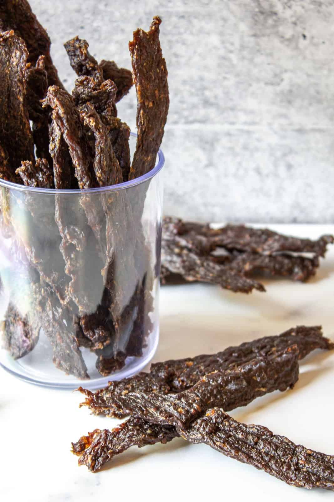 Dried jerky on a white surface with a jar filled of jerky in the background. 