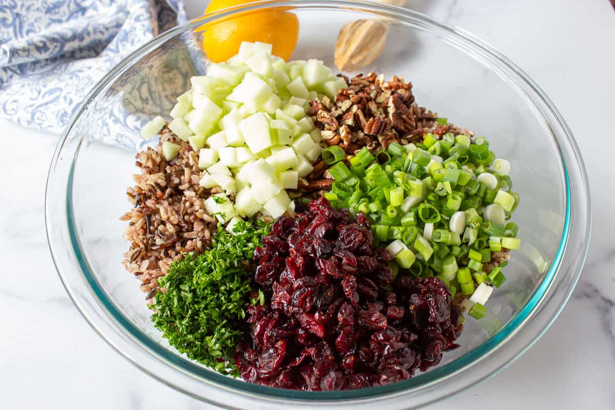 A glass bowl filled with rice, apples, cranberries, onions, pecans and parsley.