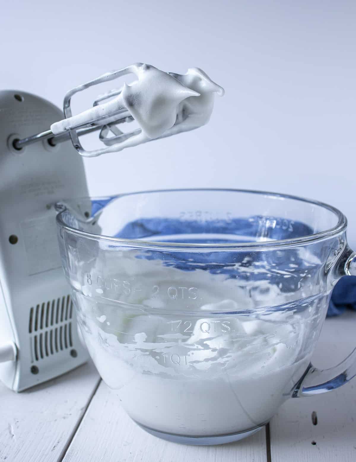A glass bowl filled with whipped egg whites next to a hand held mixer.