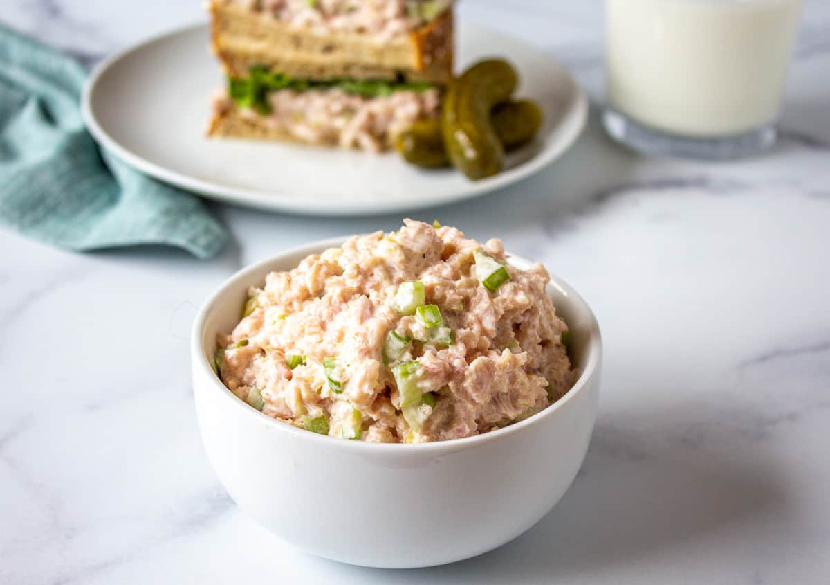 A small white bowl filled with a ham salad and a sandwich in the background. 