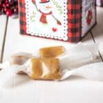Caramels wrapped in waxed paper on a white board with a snowman tin behind the caramels.