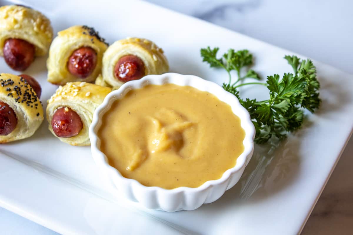 A small white bowl filled with a brown mustard surrounded by little smokie appetizers.