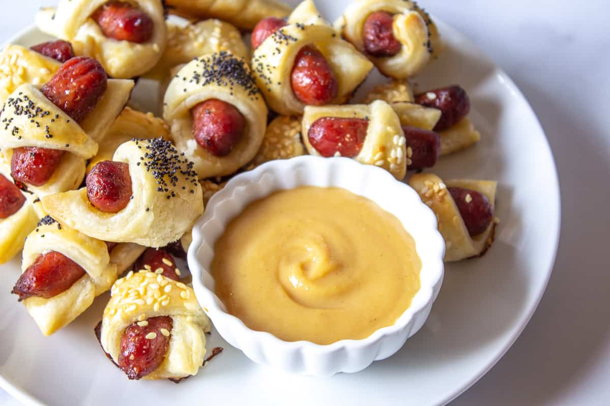 A small bowl filled with a yellow mustard surrounded by appetizer bites.