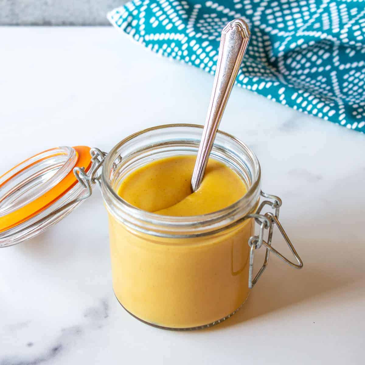 A glass crock filled with yellow mustard with a spoon in the middle of the crock.