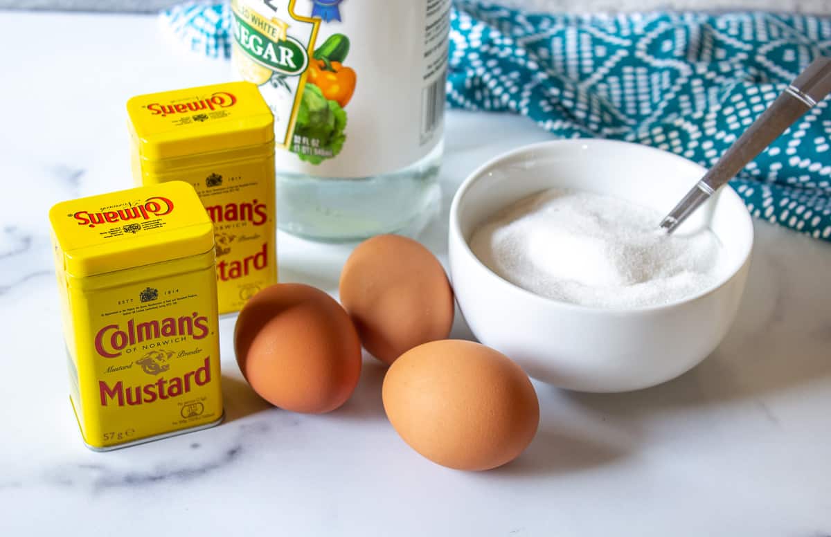 Jars of dried mustard, eggs and sugar arranged together on a white marbled surface.