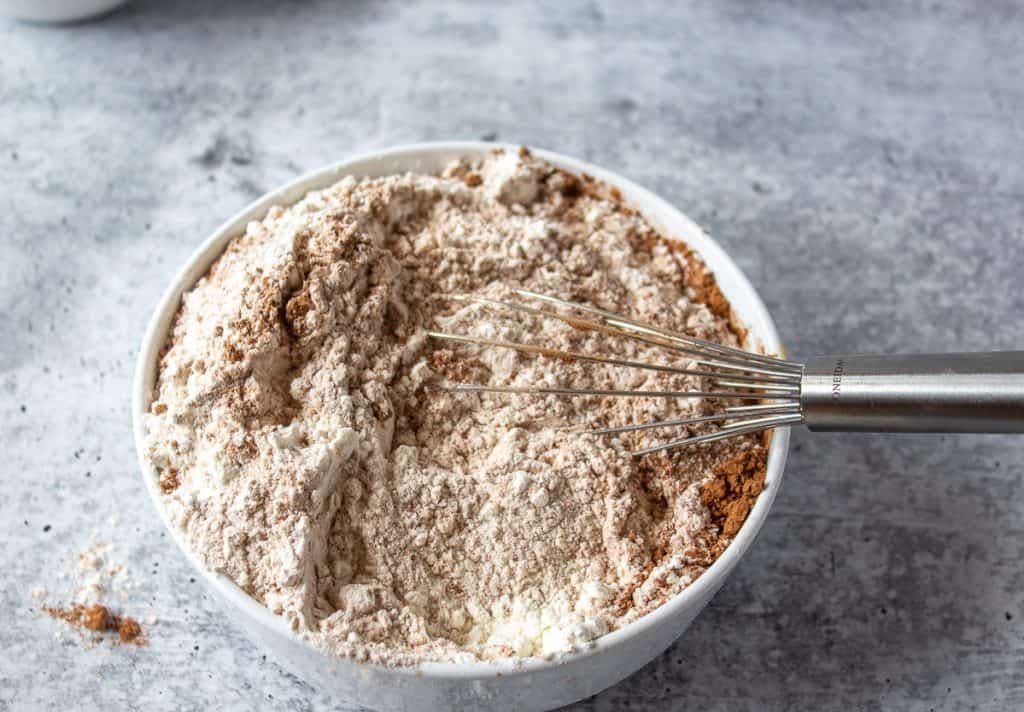 A white bowl filled with flour and cocoa powder with a wire whisk in the bowl.