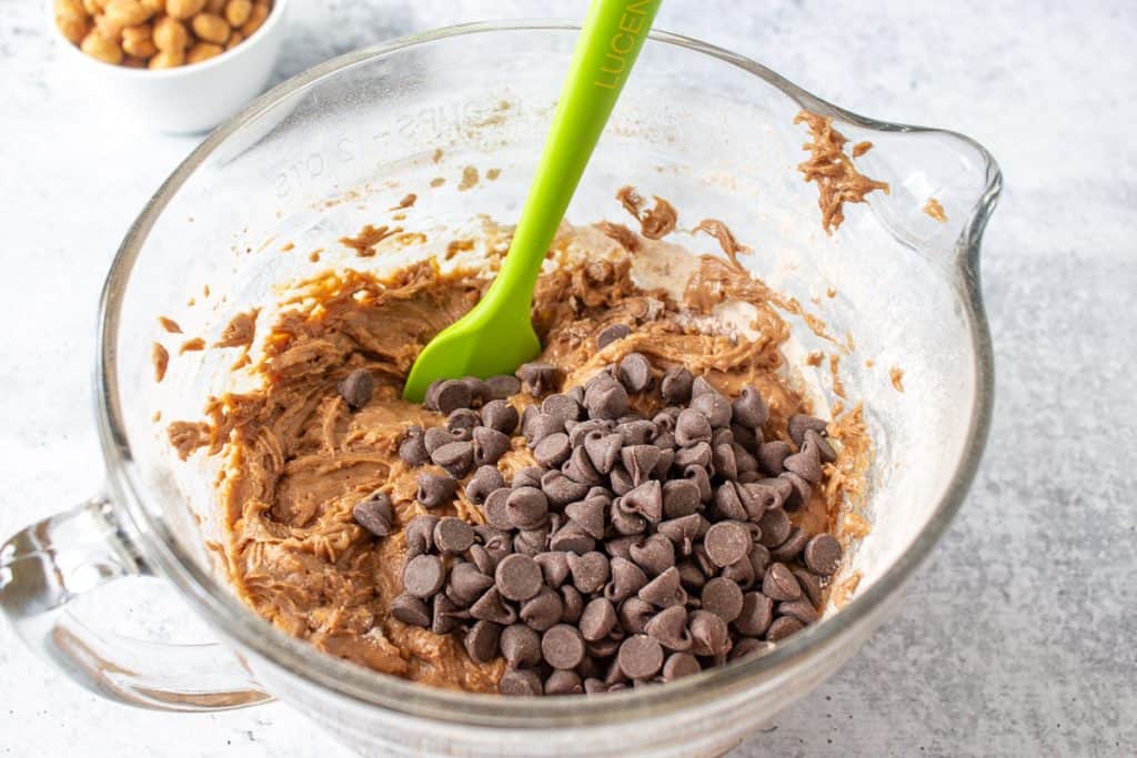 Chocolate chips on top of muffin batter in a glass bowl with a green spatula.