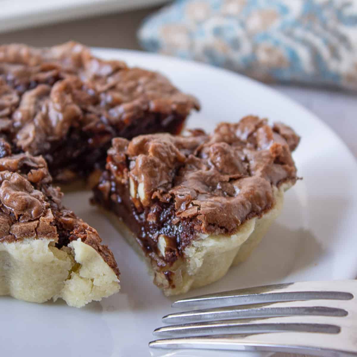 A mini chocolate tart with walnuts on a white plate with a wedge cut from the tart.