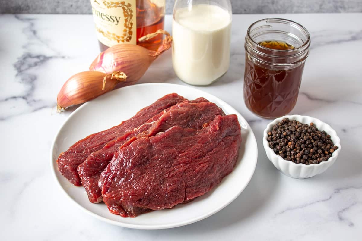 Raw elk steaks on a white plate next to a small bowl filled with peppercorns.