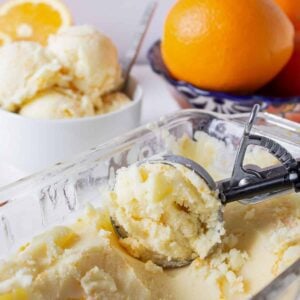 Orange ice cream in a glass dish being scooped into a round shape by an ice cream scoop.