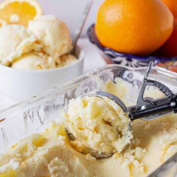 Orange ice cream in a glass dish being scooped into a round shape by an ice cream scoop.