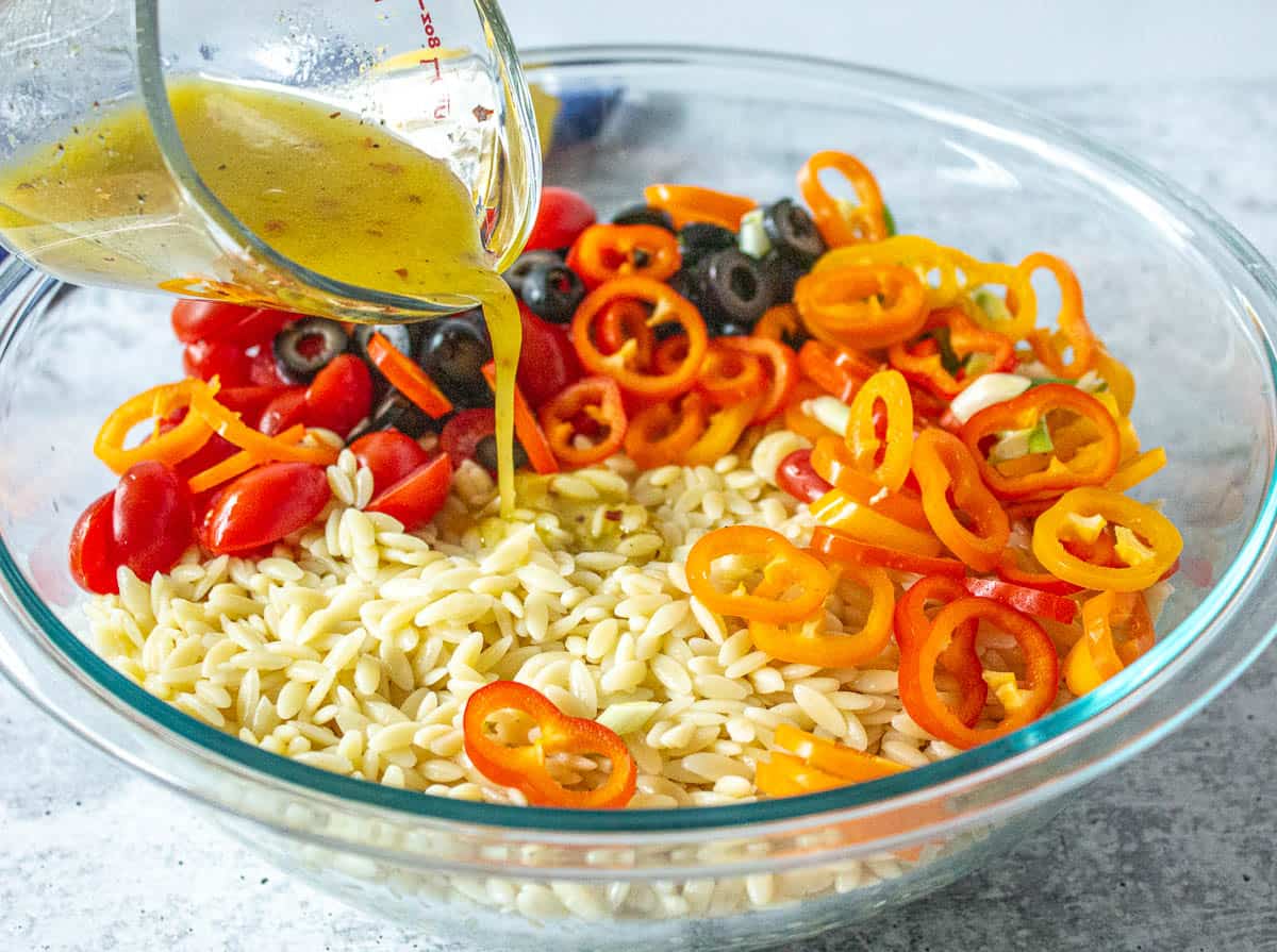 A vinaigrette being poured over a bowl filled with orzo pasta and fresh veggies.