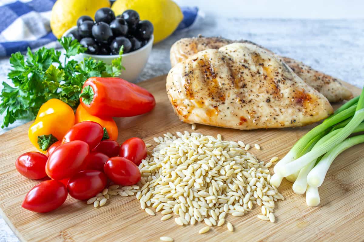 Dried orzo pasta on a cutting board along with fresh vegetables and a grilled chicken breast.