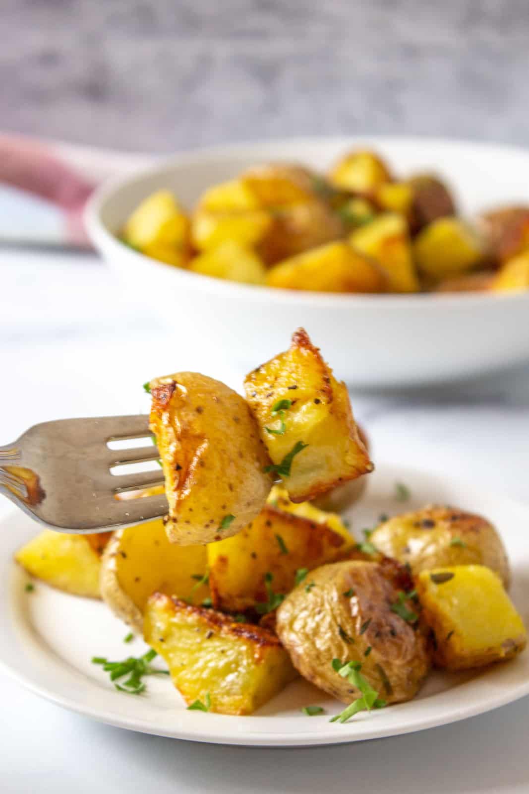 A fork with two golden roasted potatoes with a plate of potatoes in the background. 