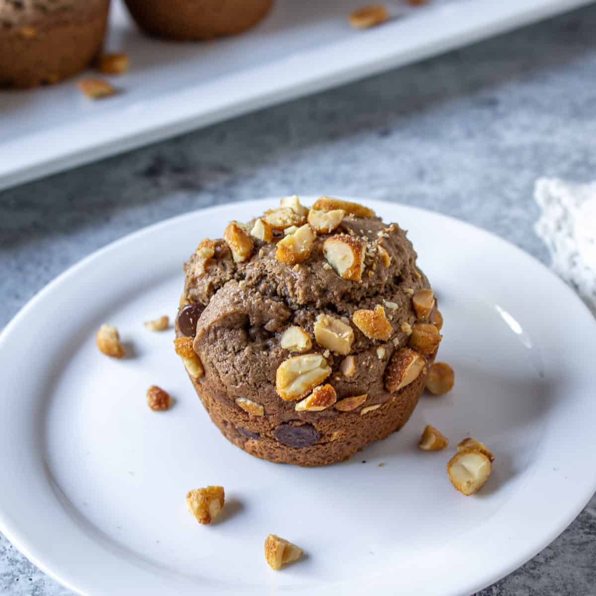 A chocolate muffin topped with peanuts on a white plate.