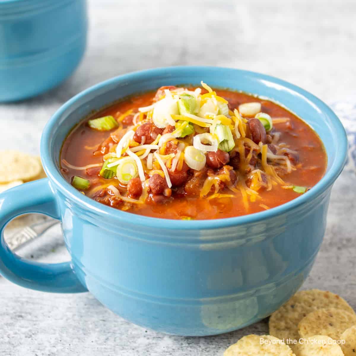 A blue mug filled with chili topped with cheese and green onions.