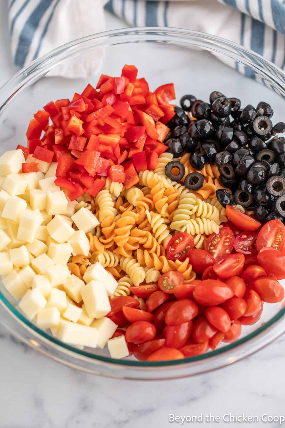 Chopped veggies, cubes of cheese, black olives and pasta in a bowl.