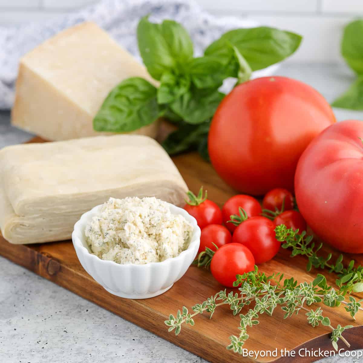 Puff pastry dough, fresh tomatoes and herbs and a soft cheese on a board. 