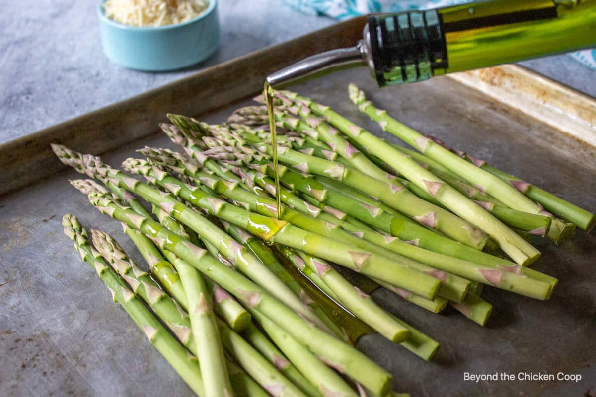 Olive oil being poured over asparagus on a baking sheet.