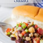 A bean salad on a plate next to a hotdog bun.