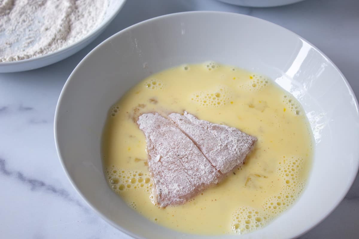 A fish fillet in a bowl with raw scrambled eggs. 