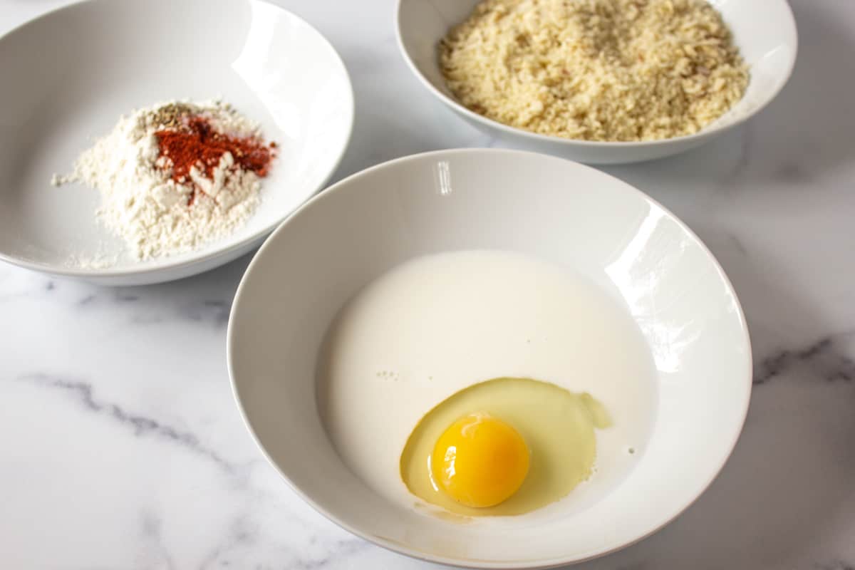 Three white bowls filled with flour, bread crumbs and egg and milk. 