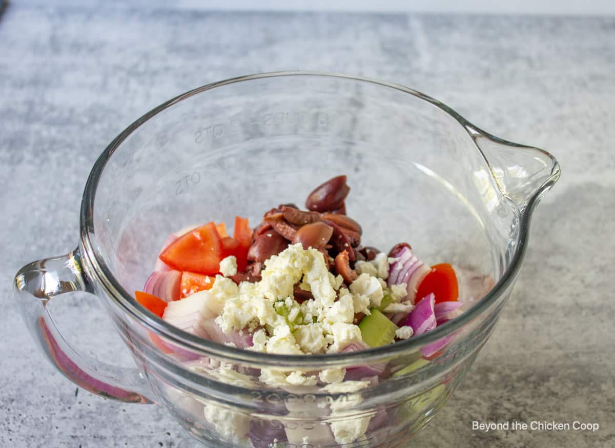 A glass bowl filled with veggies, olives and crumbled cheese.