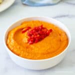 A white bowl filled with a red hummus topped with chopped red peppers.