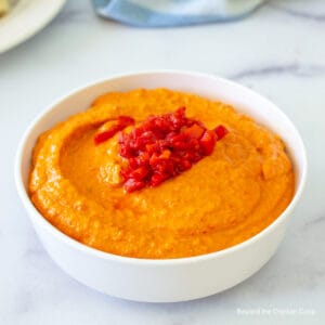 A white bowl filled with a red hummus topped with chopped red peppers.