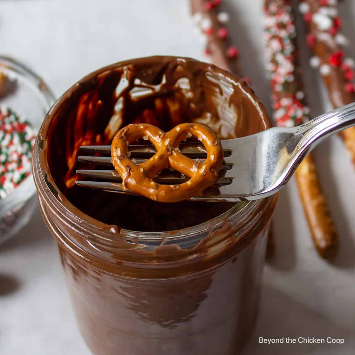 A pretzel on a fork over a jar of melted chocolate.