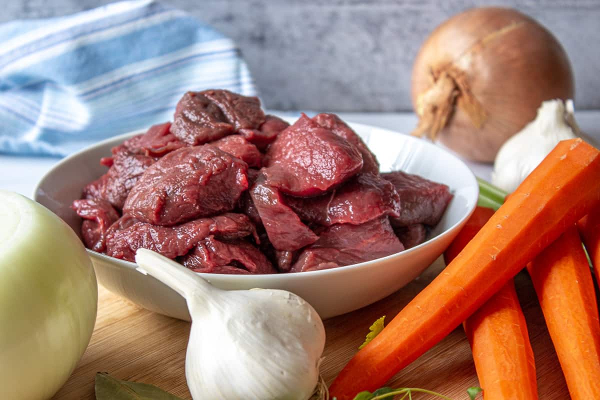 A cutting board filled with carrots, onions, garlic and cubed raw meat.