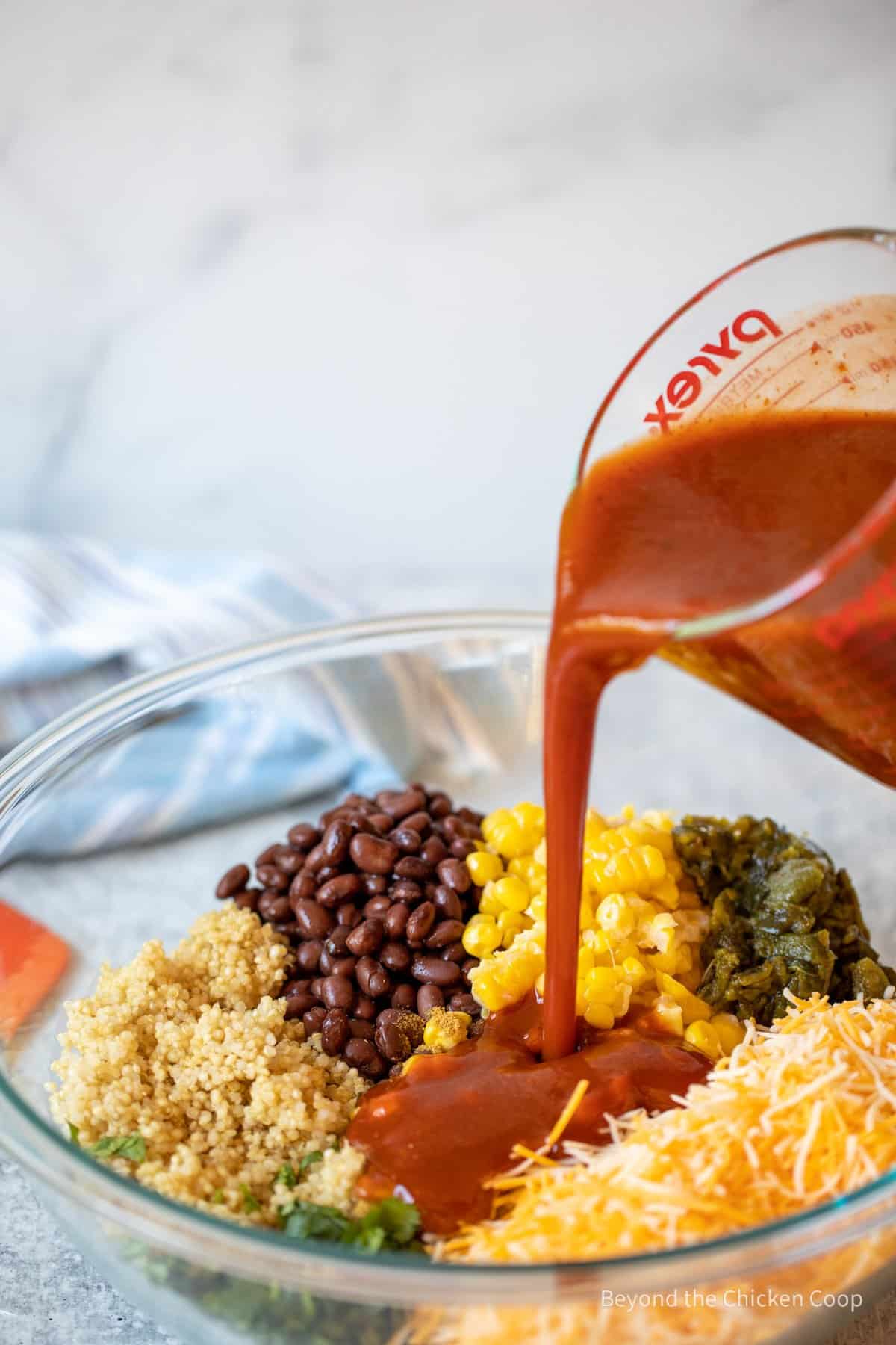 Pouring enchilada sauce over a bowl with corn, beans and cheese.